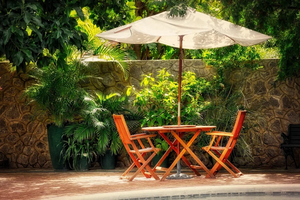 Peaceful outdoor seating area with wooden furniture, lush plants, and a stone wall backdrop. 