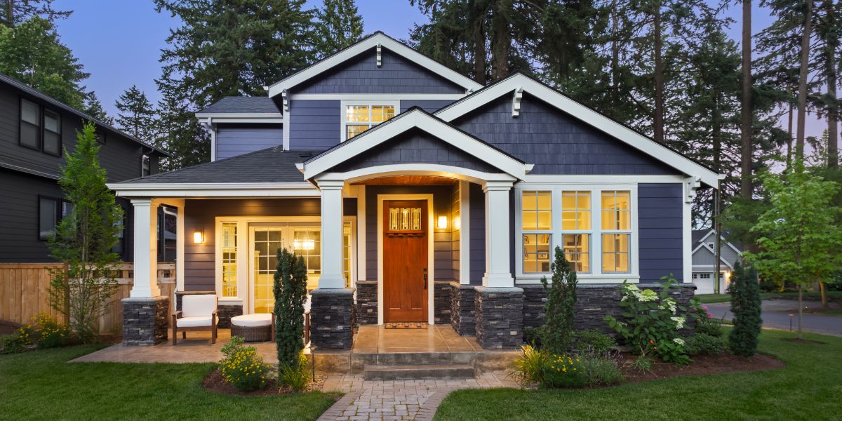 Craftsman-style home exterior with dark blue siding, white trim, stone accents, and warm wood front door at dusk.